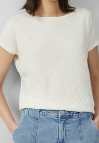 Vit texturerad blus med korta ärmar och rund halsringning, med ett subtilt veckmönster, i kombination med ljusblå denimjeans.