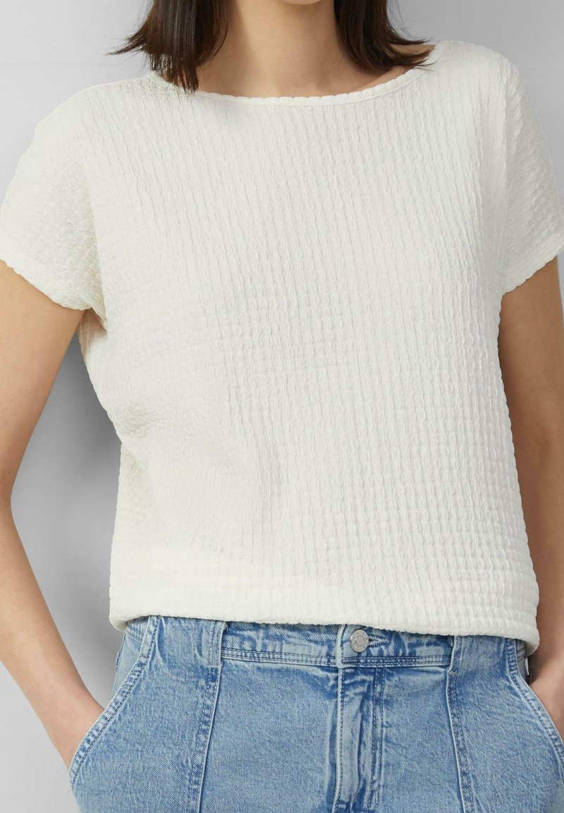 Vit texturerad blus med korta ärmar och rund halsringning, med ett subtilt veckmönster, i kombination med ljusblå denimjeans.