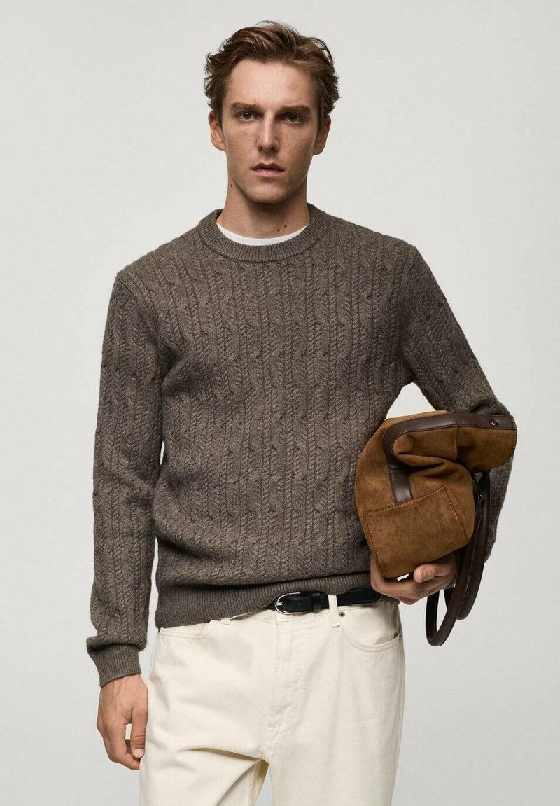 Brown cable-knit sweater with a round neckline, paired with cream-colored pants. Holding a brown suede bag with leather accents.