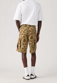 Pantalones cortos de camuflaje en caqui y marrón con múltiples bolsillos, combinados con una camiseta blanca de gran tamaño y zapatillas en blanco y negro.