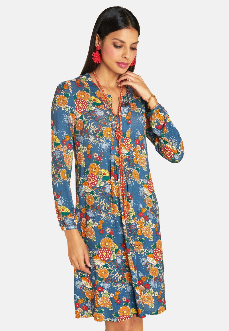Blumenkleid aus blauer Stoff mit roten, orangen und gelben Blumenmustern. Lange Ärmel, Brusttasche und eine Knopfleiste.