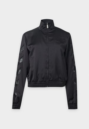Femme aux longs cheveux raides portant une veste noire zippée, une chemise blanche, un pantalon de survêtement ample foncé et des baskets noires et blanches, debout devant un fond blanc.