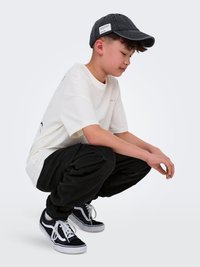 Deček, oblečen v črno kapo, belo majico, črne cargo hlače in črne teniske, se skloni z rokami sproščeno spredaj na svetlo sivi podlagi.