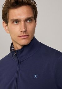 Giacca a zip navy con colletto alto, caratterizzata da un logo ricamato di colore azzurro chiaro sul petto. Tessuto liscio con una sottile foderatura a righe.