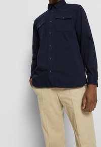 Chemise bleu marine à boutons avec deux poches poitrine et boutons-pression, associée à un pantalon beige clair. Le tissu semble lisse avec une coupe structurée.