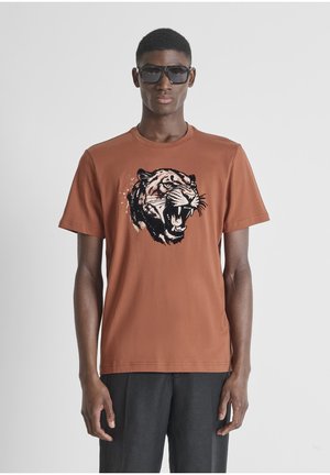 Hombre con gafas de sol negras de gran tamaño, camiseta color óxido con un gráfico de un tigre rugiente y pantalones negros, de pie frente a un fondo gris liso.