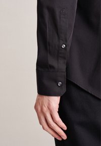 Camicia nera con bottoni, caratterizzata da una texture liscia, bottoni a contrasto e un dettaglio ricamato sul polsino. Primo piano della manica e della mano.