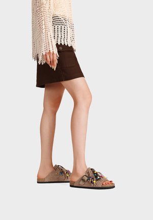 Donna che indossa sandali beige slip-on con perline colorate, gonna marrone scuro e top crema all'uncinetto, in piedi su uno sfondo semplice.