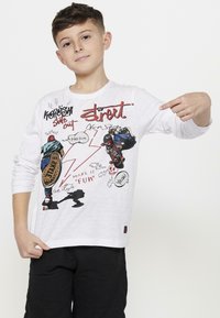 Witte lange mouwen T-shirt met kleurrijke skateboardgraphics en tekst. Het shirt heeft een relaxte pasvorm en een ronde halslijn. Zwarte broek getoond.