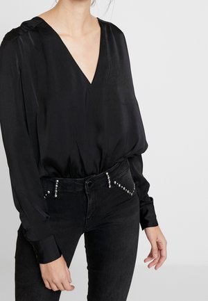 Frau trägt eine schwarze Satin-V-Ausschnitt-Bluse, die in eine schwarze Jeans mit silbernen Nieten an den Taschen gesteckt ist, und steht vor einem schlichten grauen Hintergrund.