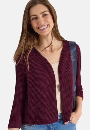 Smilende kvinde med langt brunt hår iført en tekstureret bordeaux cardigan, beige top, blå jeans og med en mørk skuldertaske.