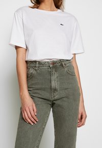 Weißes T-Shirt mit kurzen Ärmeln und grünem Lacoste-Logo, kombiniert mit olivgrünen High-Waisted-Jeans mit Knopf und fünf Taschen.
