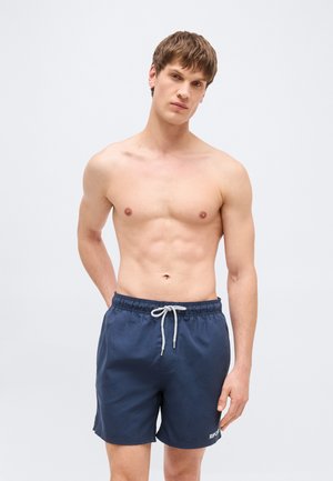 Uomo senza maglietta con capelli corti castani, indossa pantaloncini da bagno blu navy con coulisse bianca, in piedi davanti a uno sfondo chiaro e uniforme.