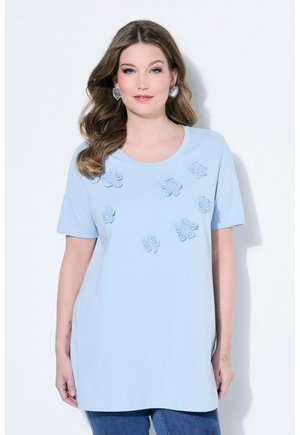 Donna con capelli lunghi e ondulati che indossa una maglietta azzurro chiaro a maniche corte con applicazioni floreali 3D e orecchini a forma di fiore argentati, in piedi davanti a uno sfondo bianco.