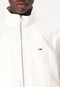 White zip-up jacket with stand collar. Lightweight, smooth texture, features a small embroidered logo in red, white, and blue on the chest.