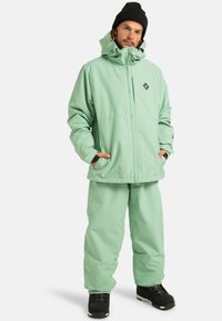 Mężczyzna w jasnoszarej zimowej kurtce i spodniach, z czarną czapką i butami, stojący z rękoma w kieszeniach kurtki na białym tle.