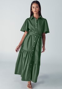 Lungo abito verde con maniche a palloncino corte, chiusura con bottoni sul davanti, vita con cintura e gonna a balze stratificate. Realizzato in tessuto morbido.