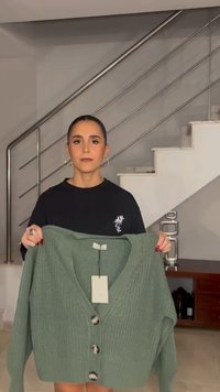 Käsipidi seljata seljatukk naine hoiab rohelise nööpidega kootud kardigani, mille küljes on hinnasilt, moodsa kodu sisekujunduse lähedal trepi kõrval.