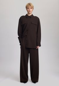 Barna, oversized ing két zsebbel és vállrész részletekkel, széles lábszárú nadrággal párosítva. Simogatú anyagból készült, matte textúrával.