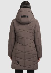 Vrouw in een lange, gewatteerde taupe jas met een capuchon en geometrische stiksels, staand met de rug naar de camera op een effen achtergrond.