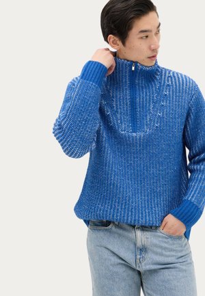 Ung mand justerer kraven på en blå tekstureret striktrøje, iført lyseblå jeans, ser mod venstre.