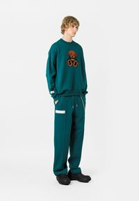 Sudadera de color aguamarina con un gráfico de oso, combinada con pantalones de chándal aguamarina a juego que tienen cremalleras laterales y un detalle de etiqueta. Calzado negro.