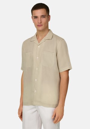 Junger Mann trägt ein beiges Kurzarmhemd mit zwei Brusttaschen und weiße Hose, steht vor einem einfarbigen Hintergrund.