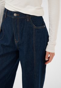 Højttaljede mørkeblå denimbukser med orange syninger, frontlommer og metalknaplukning, kombineret med en hvid, langærmet top.