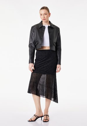 Donna con giacca di pelle nera corta, top bianco corto, gonna asimmetrica a pois neri e sandali neri con tacco e cinturini, in piedi immobile.