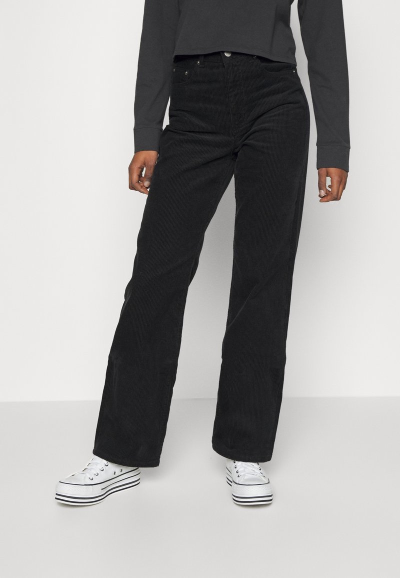 Personne portant un pantalon large taille haute en velours côtelé noir avec des baskets blanches à plateforme, debout devant un fond clair uni.