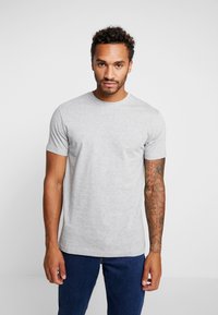 Grijze katoenen T-shirt met een ronde hals, korte mouwen en een getailleerde pasvorm. G gedragen met blauwe jeans; zichtbaar tattoeages op de armen.