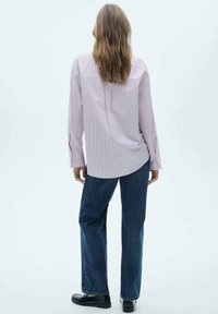 Chemise rayée rose clair à manches longues, ourlet arrondi, paire de jeans droit bleu foncé, et chaussures noires. Vue debout de dos.