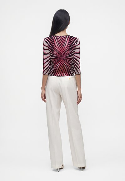 Femme aux longs cheveux noirs portant un haut rose et noir à motifs et un pantalon blanc à jambes larges, debout de dos sur fond blanc.