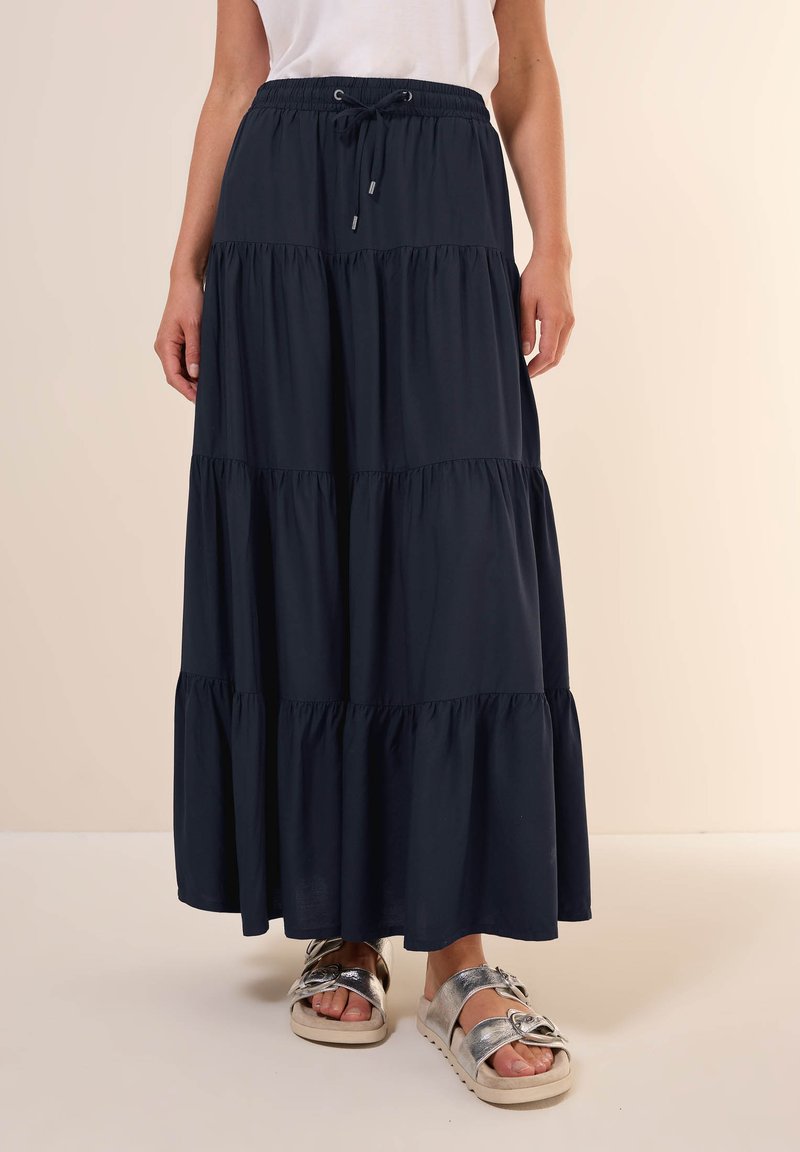 Jupe maxi bleu marine à niveaux avec une taille élastique et un cordon de serrage, fabriquée en tissu léger. Associée à des sandales argentées.