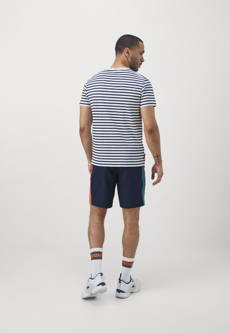 T-shirt rayé en blanc et bleu marine avec un short bleu marine à panneaux latéraux colorés. Chaussettes blanches avec des accents rouges et verts, et baskets blanches.