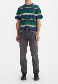 Camiseta a rayas en azul, verde, naranja y blanco; presenta un bolsillo en el pecho. Combinada con jeans grises y zapatos negros. Diseño simple y casual.