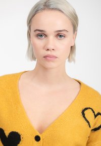 Jongeman met kort blond haar die een mosterdgele cardigan draagt met zwarte hartpatronen en een zwarte knop, neutrale uitdrukking.