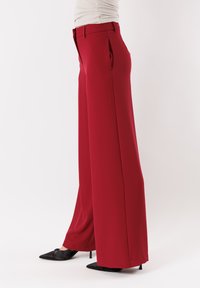 Pantaloni rossi a gamba larga con una trama liscia, caratterizzati da pieghe frontali e una vita sartoriale. Abbinati a tacchi neri a punta.