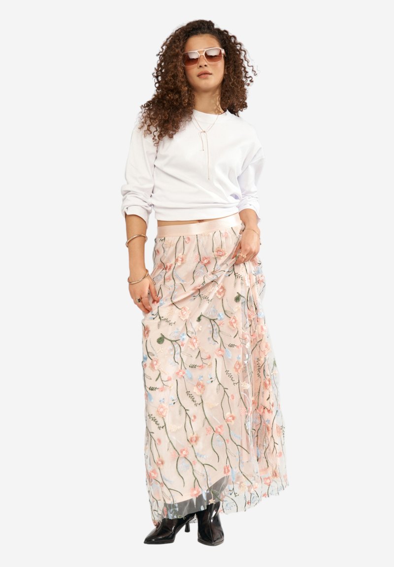 Femme aux cheveux bouclés portant des lunettes de soleil couleur sable, un sweat-shirt court blanc, une jupe longue brodée de fleurs, des bottines noires et des bijoux en or.