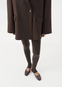 Cappotto oversize marrone con rever a scaldabraga, abbinato a collant a coste scuri e ballerine bordeaux. Texture liscia e design sartoriale.