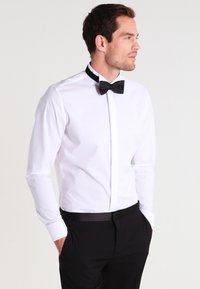 Camicia bianca con colletto classico, abbinata a un papillon nero e pantaloni neri. La camicia ha un design aderente e polsini.