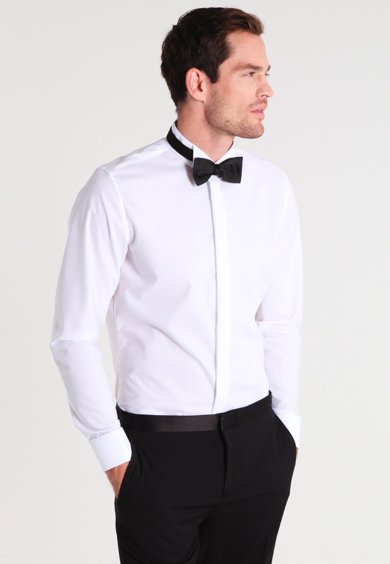 Man draagt een wit overhemd met een zwarte strikdas en zwarte broek, staat met zijn handen in zijn zakken, kijkt naar rechts.