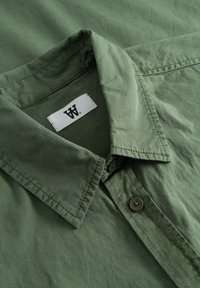 Camicia verde con bottoni, realizzata in un tessuto morbido e testurizzato, con colletto pieghevole e un'etichetta con logo. Bottoni e dettagli di cucitura visibili.