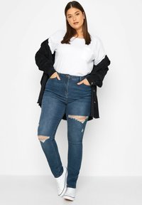 Jeans de tiro alto en azul oscuro con rodillas desgastadas, combinados con una camiseta blanca y una chaqueta negra, y zapatillas blancas.