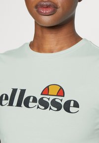 Camiseta de algodón verde menta con un prominente logo negro de "ellesse" acompañado de un acento semicircular rojo y amarillo en la parte superior. Mangas cortas, escote redondo.