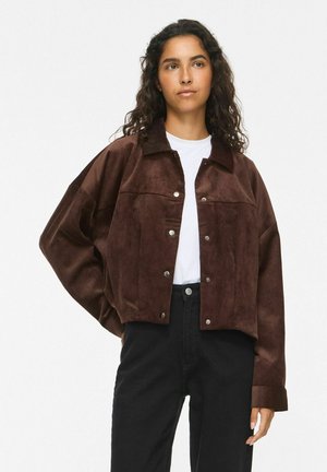 Femme aux longs cheveux bouclés portant une veste en daim marron à boutons par-dessus une chemise blanche et un pantalon noir taille haute, debout devant un fond uni.