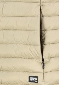 Beige gesteppter Stoff mit horizontalen Nähten, teilweise offener schwarzer Reißverschluss und einem schwarzen rechteckigen Etikett mit der Aufschrift "Original 86 Always Be Bold Sublevel."