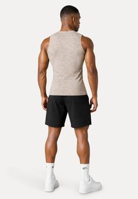 Homme musclé de dos portant un débardeur beige, un short noir, des chaussettes blanches et des baskets blanches, debout sur un fond uni.