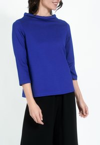 K�ék, hosszú ujjú blúz, cseppnyakú; puha anyagból készült; laza fazonnal és egyenes alsó szegéllyel. Fekete nadrággal viselet.