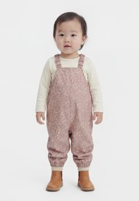 Bambino in piedi rivolto in avanti, che indossa una maglietta chiara a maniche lunghe, salopette rosa con motivi floreali e stivaletti beige alla caviglia su sfondo bianco.